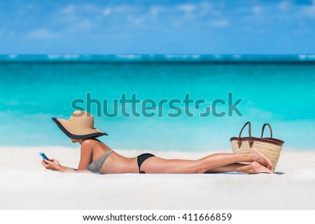 Beach vacation girl using mobile phone app texting sms or sharing photos on social media during summer travel holiday. Bikini woman relaxing sunbathing on sand lying on towel wearing sun hat.