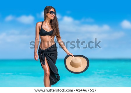 Elegant beach woman in bikini and fashion sarong standing on shore. Sexy lady in black beachwear, floppy hat, sunglasses enjoying sun on tropical destination during summer vacation in the Caribbean.