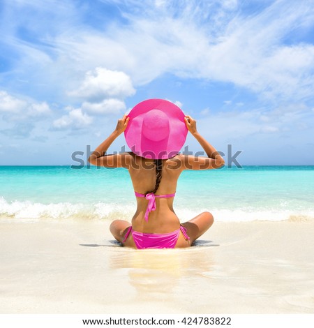 Perfect paradise summer vacation happiness carefree happy woman relaxing sitting in sand enjoying tropical beach destination. Back view of bikini girl holding pink straw hat on Caribbean holiday.