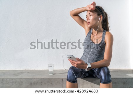 Tired fitness woman sweating taking a break listening to music on phone after difficult training. Exhausted Asian runner dehydrated feeling exhaustion and dehydration from working out at gym.