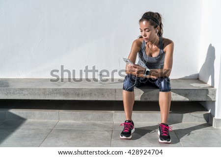 Young woman listening to music with earphones on smart phone app for fitness motivation. Athlete runner in sportswear relaxing sitting getting inspired. Asian mixed race model.