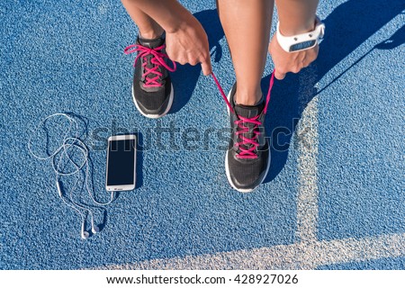 Runner woman tying running shoes laces getting ready for race on run track with smartphone and earphones for music listening on mobile phone. Athlete preparing for cardio training. Feet on ground.