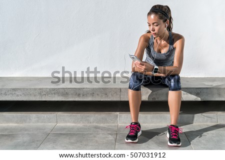 Fitness runner on mobile smart phone app tracking progress listening to music with earphones for fitness motivation. Athlete runner in sportswear relaxing sitting getting inspired. Asian mixed race.