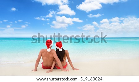 Christmas beach vacation holidays santa hat couple relaxing from behind sitting on white sand wide horizontal sky background for text advert for holiday season. Blue ocean texture.