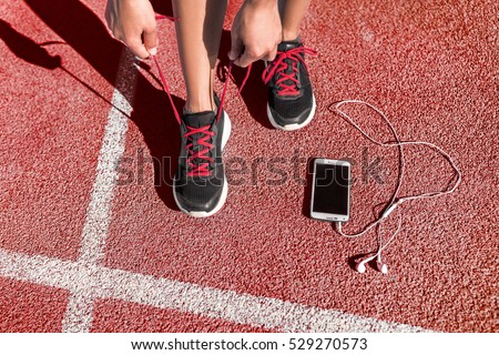 Runner woman athlete tying running shoes laces getting ready for race training on run track with smartphone and earphones for music listening on mobile phone.