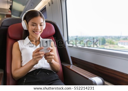 Modern people city lifestyle. Young urban woman using phone app and wireless headphones to listen to music or play video games online. Asian girl enjoying train travel in business class seat.