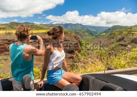 Road trip vacation car couple taking pictures of Hawaii nature landscape with smartphone camera app. Young people relaxing in convertible car on summer holidays. Travel lifestyle.