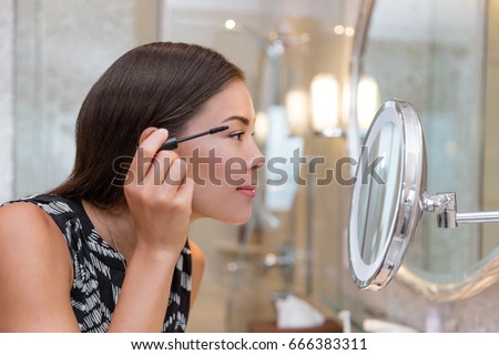 Woman getting ready for work doing morning makeup routine putting mascara in bathroom mirror at home. Beautiful Asian businesswoman applying eye make-up.