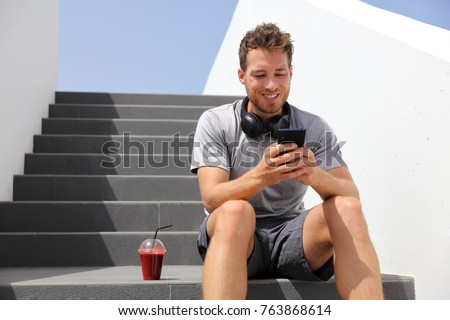 Healthy active lifestyle man using smart mobile phone app while drinking red smoothie holding cellphone on jogging break with morning beet juice sitting on stairs outdoor.