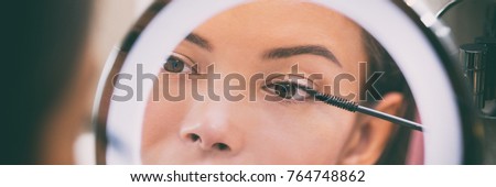 Woman putting mascara makeup in mirror banner at home bathroom morning routine. Beautiful Asian girl getting ready applying eye make-up with brush. Closeup on eye in reflection.