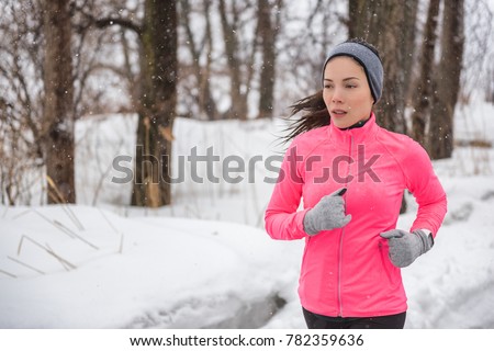 Winter sport fitness girl running in snow wearing wind jacket, gloves, headband and smartwatch. Asian woman healthy and active lifestyle training outside during the cold weather winter season.