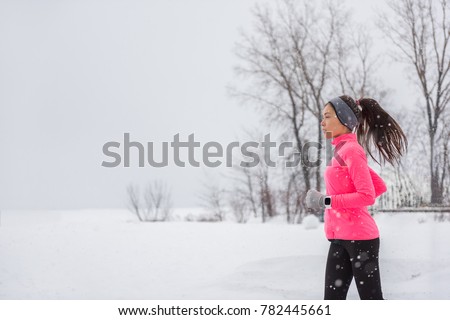 Winter running woman in cold snow weather jogging outside wearing windproof clothes with gloves, headband, winter tights and wind jacket in white snowing storm background.