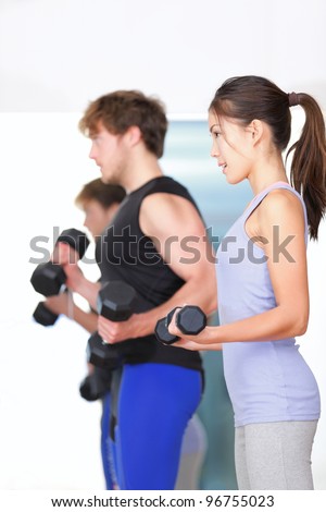 Fitness people in gym. Couple strength training lifting weights during indoor fitness workout. Woman lifting dumbbells training biceps in focus.