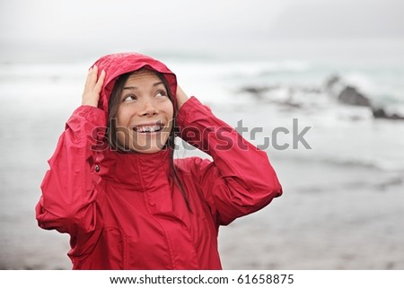 Woman smiling at rain in red raincoat on the beach on a cold raining fall day. Caucasian / Asian woman.