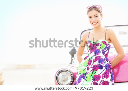 Car woman smiling happy standing in front of pink retro vintage car. Portrait of pretty girl in summer dress.  Multicultural Chinese Asian / Caucasian female lifestyle model. Photo from Havana, Cuba.