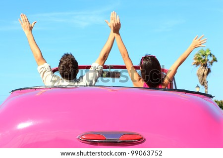 Freedom - happy free couple in car driving in pink vintage retro car cheering joyful with arms raised. Friends going on road trip travel on summer day under sun blue sky.