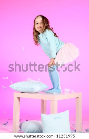 Five years old girl having fun with blue pillows and feathers at small table on pink colored background.