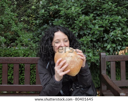 Portrait of hungry pretty young goth brunette girl woman female model eating huge bread seated on bench in a London park, UK