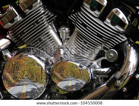 Close up of a twin cylinder motorcycle engine with reflection of fall colored tree lined highway.