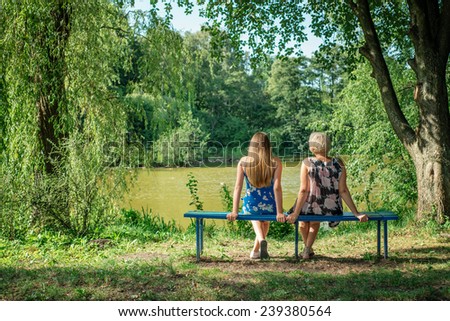 Two women of different generations sitting on a bench near a pond in the summer. Mother and daughter hugging. Grandmother and granddaughter.