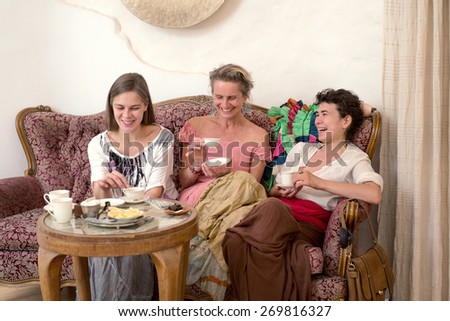 Three women in ethnic dresses sitting on an old couch and drinking tea