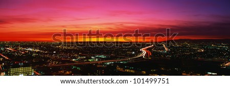 This is an aerial view of downtown Los Angeles at sunset. The streaked lights of the freeway are in the center with an orange sunset sky.