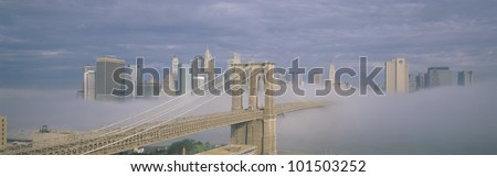 This is the Brooklyn Bridge over the East River with the Manhattan skyline without the World Trade Center behind it. There is a morning fog enveloping the bridge.