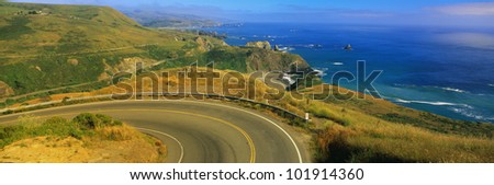 This is the Pacific Coast Highway. The road curves around a bend to the left and drops down overlooking the ocean. The rocky hillside is also seen next to the ocean.