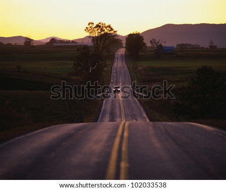 This is a country road at sunset in the Adirondack area. The road trails off into infinity. There is a car coming forward.
