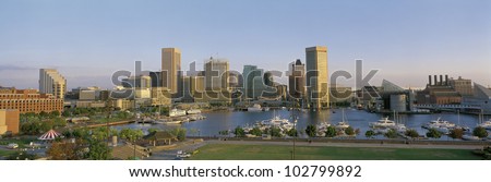 This is the skyline of Baltimore in daylight showing the inner harbor and city lights. There are boats moored in the harbor
