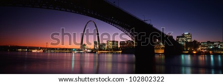 This is the St. Louis skyline and Arch at sunset. Above it is the Eads Bridge along the Mississippi River. There is a purple cast in the sunset sky.