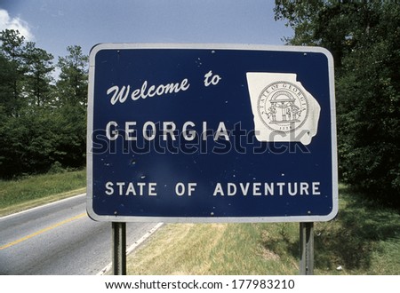 This is a road sign that says, welcome to Georgia, state of adventure. There is a white map of the state of Georgia on its right. There are green bushes on the sign\'s right and left.