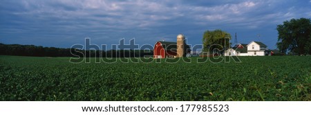This is a farm with a silo and barn. Directly behind it sits a white farmhouse. It sits in the middle of a green farm field.