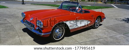 This is a restored 1959 Corvette. It is bright red with a white side panel with white sidewall tires. The convertible top is down. It is parked on flat pavement.