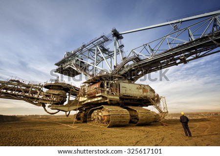 Coal mine worker with a helmet on his head is standing in front of huge drill machine and looking at it. Rear view.