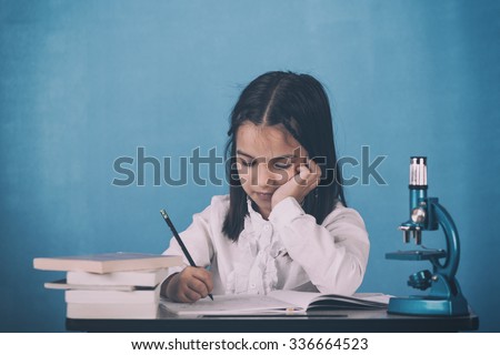 Color, old fashioned image of a serious, little girl student sitting at a school desk, thinking and writing. Blue background behind. Photo is carefully post processed for achieving finest film look.