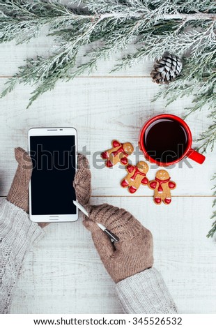 Vintage winter background of woman hands in gloves holding cellular phone. Cup of Tea and gingerbread cookies on white wooden table.
