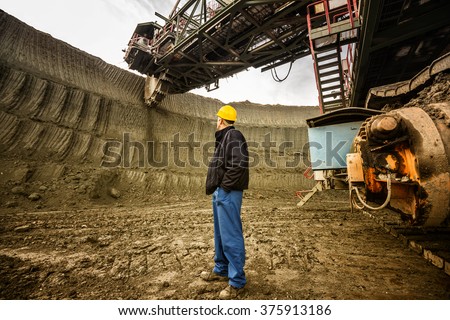 Coal mine worker with a helmet on his head standing in front of huge drill machine and looking at it. Rear view.