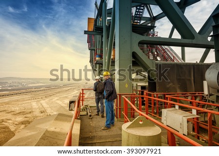 Two coal mine engineers with protective helmets standing on a huge drill machine, talking and watching at the digging site. Beautiful and colorful sky in the background. Rear view.