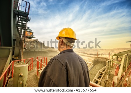 Coal mine engineer with a helmet standing on a huge drill machine and looking at digging site. Rear view. Beautiful and colorful sky in background.
