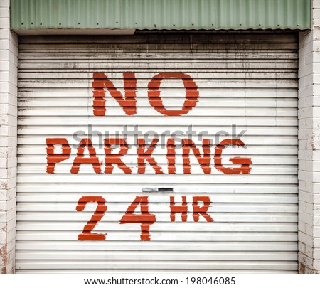 Hand-painted red sign on grimy cream colored lane garage roller door stating NO PARKING 24 HR