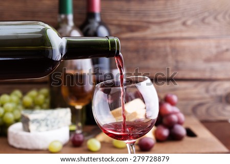 Red wine pouring into wine glass, close-up. ?heese, grapes and wine bottles on wooden table in restaurant. Flat mock up for design