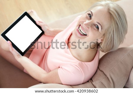 Smiling beautiful middle aged woman sitting on couch with tablet white s?reen and looking up. Unusually top view