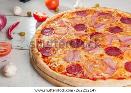 Close up Delicious Meat Pizza with Salami, Bacon and Tomato Slices on a cutting board on white wooden background with different colourful vegetables