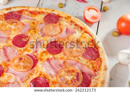 Close up Delicious Meat Pizza with Salami, Bacon and Tomato Slices on a cutting board on white wooden background with different colourful vegetables top view