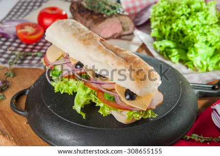 Fresh sandwich with wheat bun, ham, bacon, cheese and vegetables on inverted pan on the table with colourfull ingredients