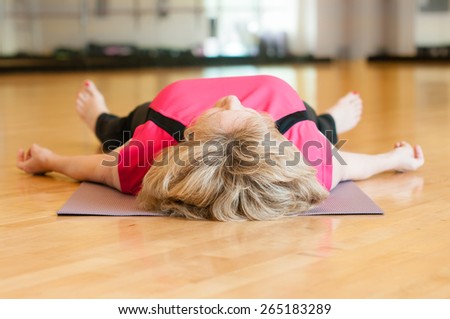 An older woman in a pink yoga outfit rests on the floor of the gym room in savasana (corpse) pose after yoga class,