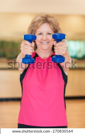 An attractive older woman smiles as she holds hand weights straight out towards the camera in an exercise studio.  The plastic-coated blue dumbbells are in sharp focus, and the woman is in background.