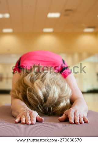An unusual angle of an older woman resting her forehead on her yoga mat in child\'s pose.  The blond woman is wearing a bright pink shirt and she is practicing on a mauve colored yoga mat.