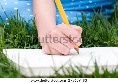 A child's hand holds a yellow pencil and draws in a sketchbook.  The child is sitting in the grass and her blue dress is visible in the background